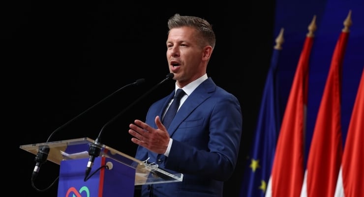 Man in a blue suit delivering a speech at a lectern with microphones, flags in the backgrounded stage setting.