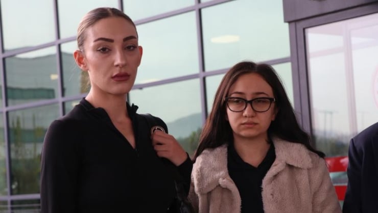 Two women stand indoors by a glass wall, looking serious; one in a black top with a ponytail, the other in a beige coat and glasses.